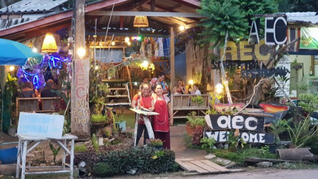 Two smiling people in red aprons stand outside a colorful, lively restaurant with AEC Welcome signs and outdoor lights.