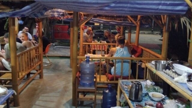 People sit and dine in a bamboo outdoor restaurant at night; a water dispenser and kitchen items are in the foreground.