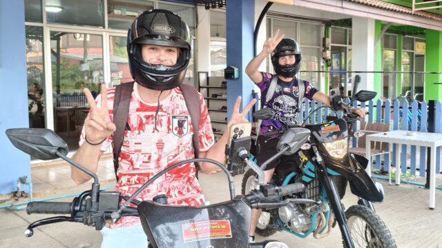 Two people wearing helmets sit on motorcycles, smiling and making peace signs in front of a colorful building.