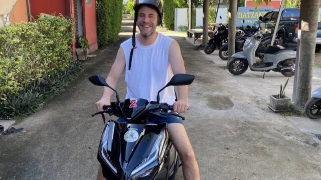 A man in a helmet and sleeveless shirt smiles while sitting on a black scooter, surrounded by palm trees and parked scooters.