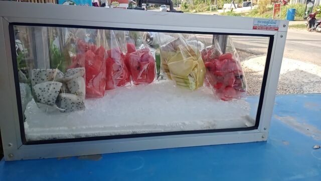 Sliced fruits in plastic bags, including dragon fruit, watermelon, and melon, displayed on ice in a glass case at an outdoor stand.