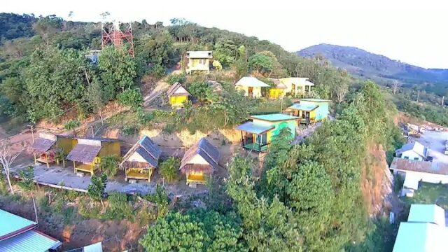 Wooden cabins and modern houses on a green hillside, surrounded by trees, with mountains in the background.