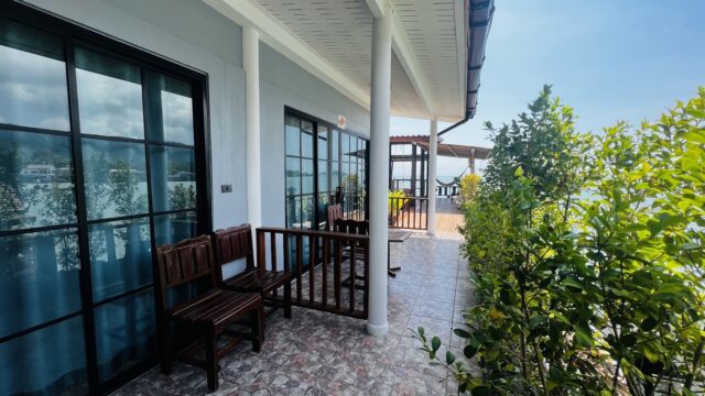A tiled balcony with wooden chairs and green plants, overlooking a sunny outdoor area with blue skies.