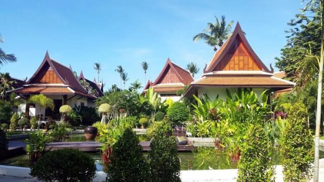 Traditional Thai-style houses with steep roofs are surrounded by lush gardens and a pond under a clear blue sky.