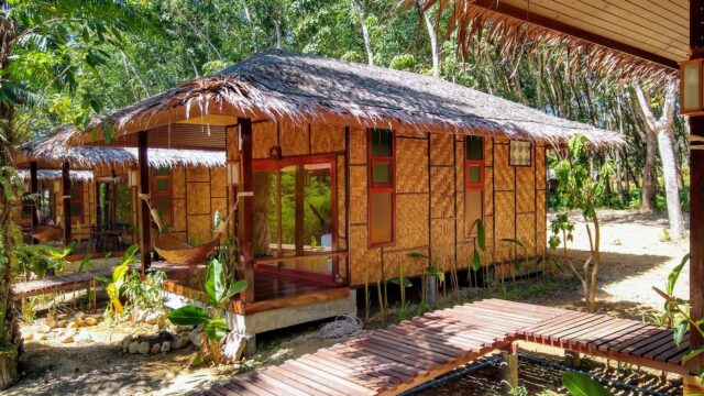 A small bamboo cottage with a thatched roof, surrounded by trees, connected by wooden walkways in a tropical setting.