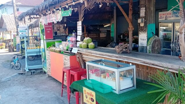Outdoor street food stall with coconuts, snacks in a display case, and colorful signs, set in a rustic, tropical setting.