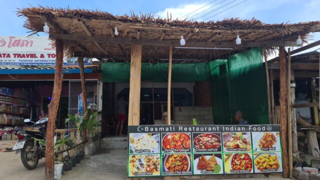 Small rustic restaurant with a thatched roof and a large menu board displaying Indian dishes outside the entrance.