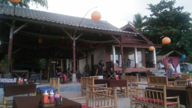 Outdoor beachside restaurant with bamboo chairs, brown tablecloths, hanging lights, and a few people near a wooden building.