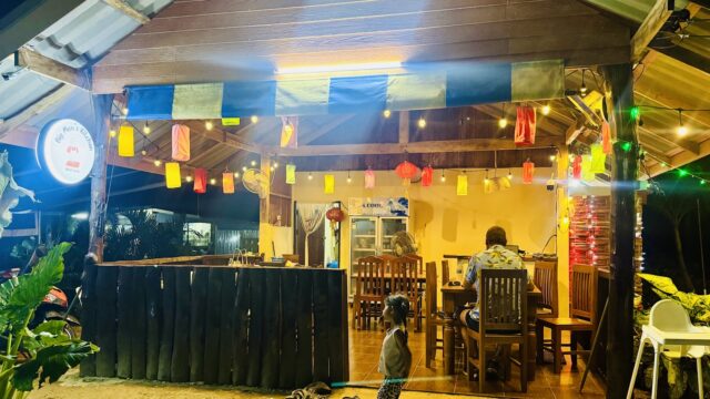 A child stands outside a brightly lit wooden restaurant with hanging lanterns and people dining inside.