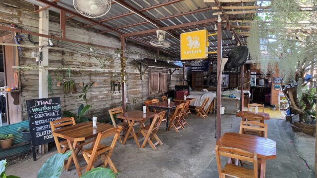 Outdoor cafe with wooden tables and chairs, plants, a Singha beer sign, and a chalkboard Thai food menu under a metal roof.