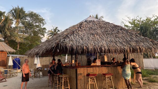 People gather at a tropical beach bar with a thatched roof, surrounded by palm trees and sand.