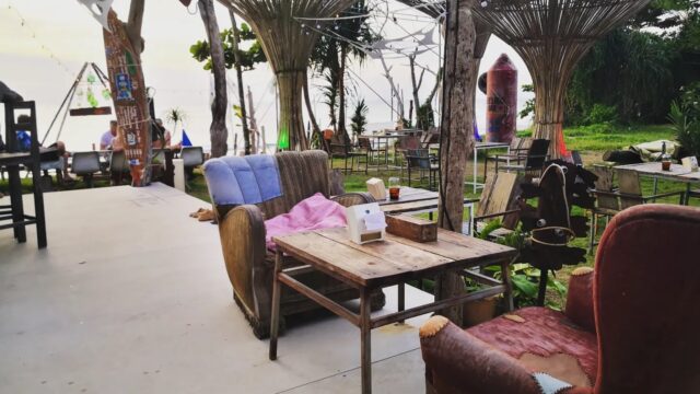 Outdoor café with rustic wooden tables, mismatched chairs, and unique bamboo decor, overlooking greenery and the sea in the background.