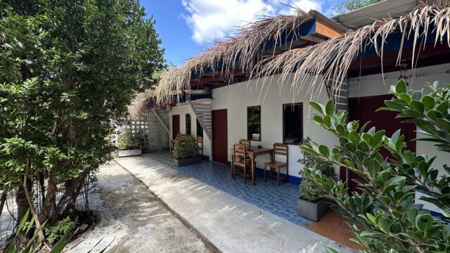 A small white building with a thatched roof, blue tiles, wooden doors, and outdoor seating, surrounded by green plants under a bright sky.