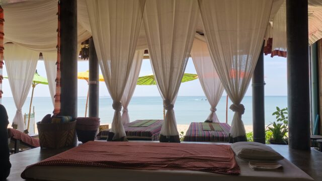 A seaside spa with beds, white curtains, and umbrellas, overlooking a calm ocean under a bright sky.