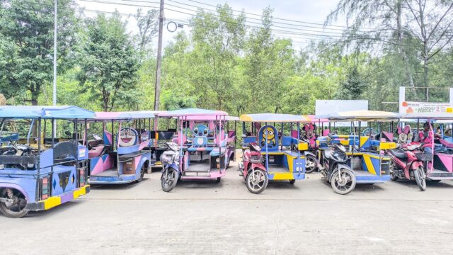 A row of colorful tuk-tuks and motorbikes parked on a street with trees and power lines in the background.