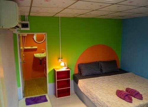 Colorful room with a double bed, two purple towels, vibrant green and orange walls, and an open door to a bathroom.