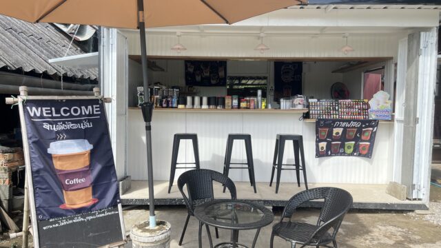 Outdoor coffee shop with a table, chairs, a large umbrella, a welcome sign, and a white counter with drinks and stools.