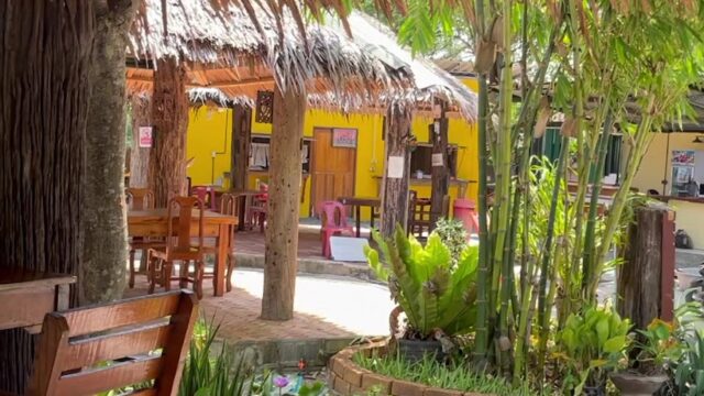 Outdoor seating under a thatched roof, facing a courtyard with plants and yellow buildings in the background.