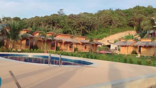 A round swimming pool in front of wooden huts with thatched roofs and lush green trees in the background.