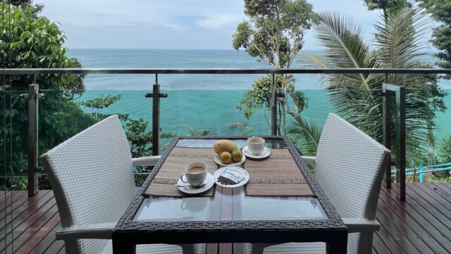 Two white chairs and a table set for two on a balcony overlook a turquoise ocean and lush green trees.