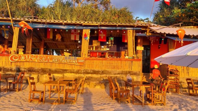 Beachfront restaurant with wooden tables on sand, colorful flags, staff in red, and warm evening sunlight.