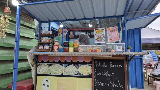 A small food stall with colorful ingredients, a menu board listing shakes, and a person working behind the counter.