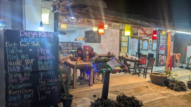 Outdoor Thai restaurant at night with diners, hanging lights, chalkboard menu, and colorful decor.