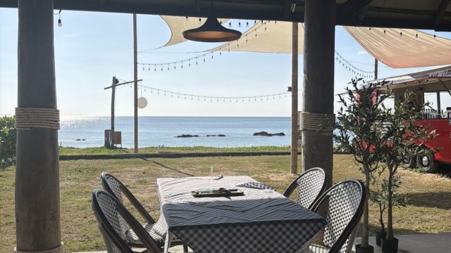 Outdoor restaurant with checkered tables, chairs, string lights, and ocean view under a sunny sky.