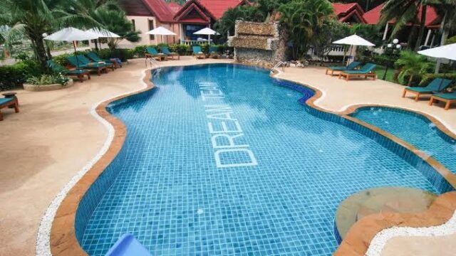 A clear blue pool with DREAM TEAM on the floor, surrounded by lounge chairs, umbrellas, and tropical plants near villas.