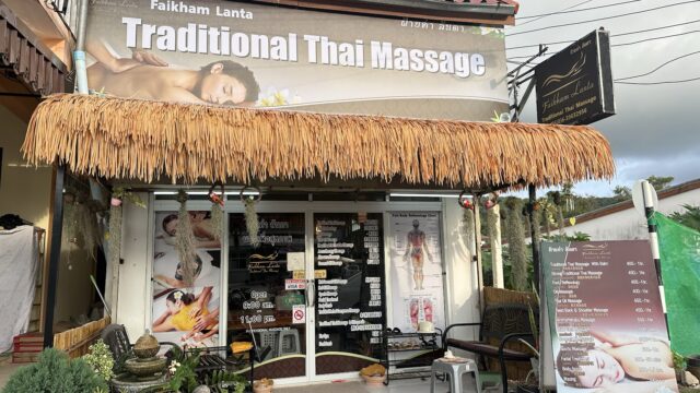 Traditional Thai massage shop with a thatched awning, sign, and price menu outside; glass doors display service ads.
