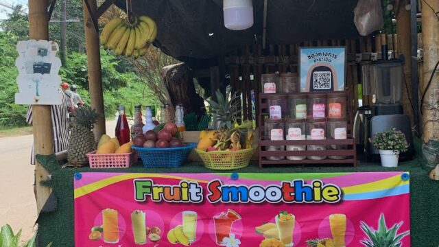 A small bamboo smoothie stand with colorful fruit smoothie menu, fresh fruit, and a blender displayed under a thatched roof.