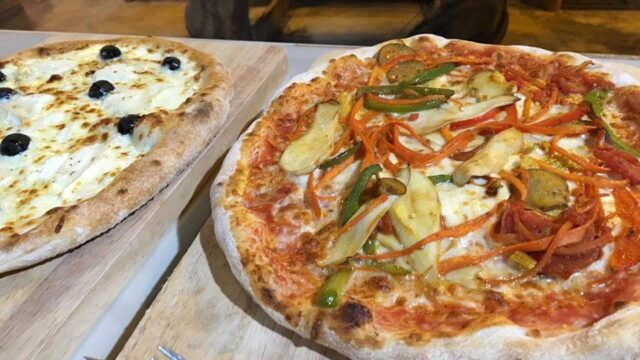 Two wood-fired pizzas on wooden boards, one with veggies, the other with cheese and black olives, on a table with forks nearby.