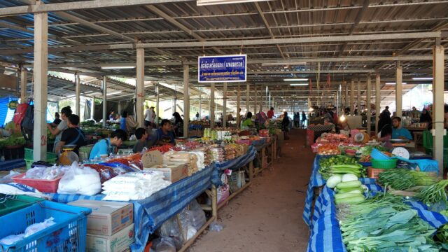 Covered market with people shopping for fresh vegetables, fruits, and packaged goods on tables under a metal roof.