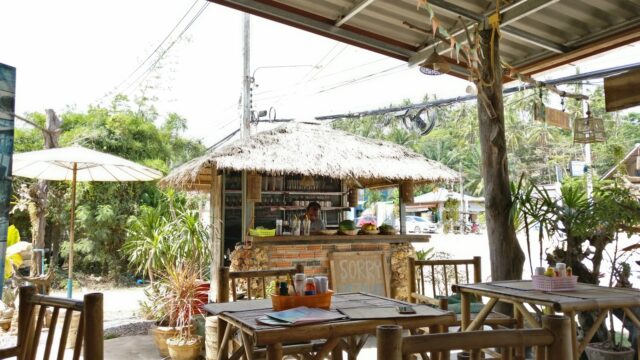 Open-air cafe with bamboo furniture, a thatched-roof counter, plants, and sunlight streaming in from outside.
