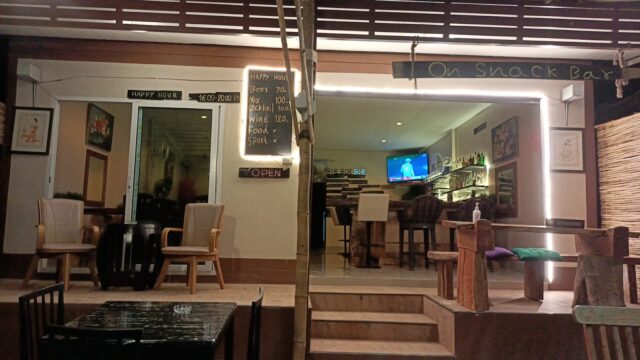 Outdoor view of a cozy snack bar at night, with tables, chairs, chalkboard menus, and a TV inside showing a sports game.