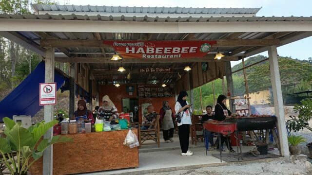 Open-air restaurant with women cooking and serving food, sign reads Habeebee Restaurant, surrounded by plants and greenery.