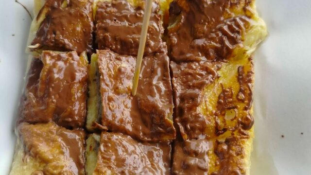 Sliced roti topped with melted chocolate, served on white paper with a wooden skewer.