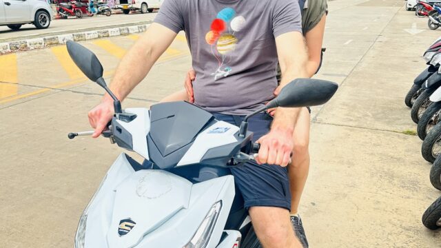 Two people in helmets sit on a white scooter parked on a street with shops and motorcycles in the background.