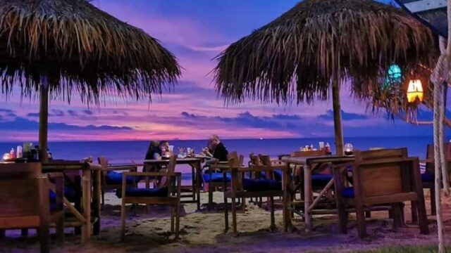 Two people dine at wooden tables under thatched umbrellas on a beach at sunset, with vibrant purple and pink skies.