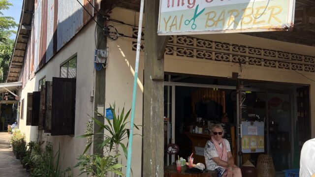 A woman sits outside Yais Barber shop, surrounded by potted plants on a sunny day.