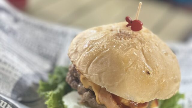 A cheeseburger with lettuce and tomato on a bun, served on newspaper-style paper, with a drink in the background.