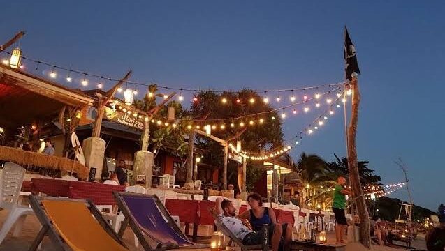 People relax on a beach at sunset, surrounded by string lights, colorful chairs, tables, and a nearby open-air restaurant.