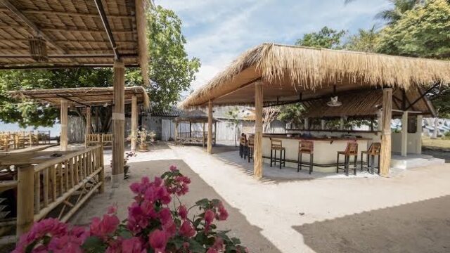 Thatched-roof outdoor bar and seating area on a sunny day, with pink flowers and trees surrounding the space.