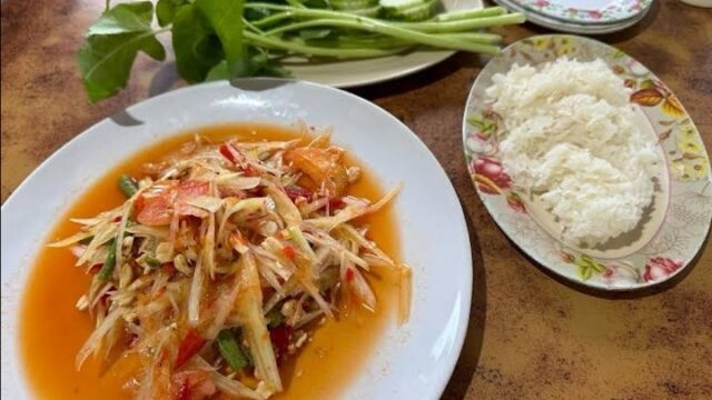 Plate of Thai papaya salad with tomatoes and chili, served with white rice, cucumber slices, and fresh greens on the side.