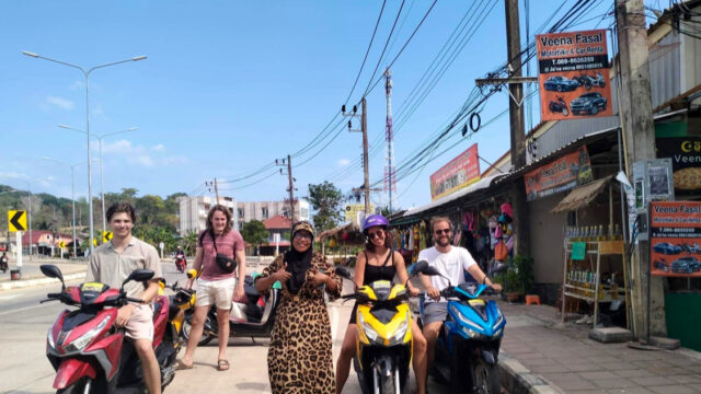 Five people pose with scooters on a sunny street lined with shops, signs, and utility poles in an urban area.