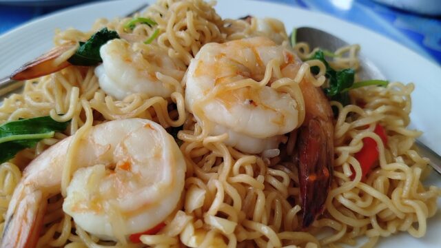 A plate of noodles with shrimp, vegetables, and a spoon, with another dish and sliced cucumbers in the background.