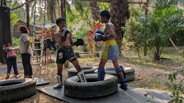 Two boys in boxing gear spar outdoors on mats and tires, with others watching in a lush, tropical setting.