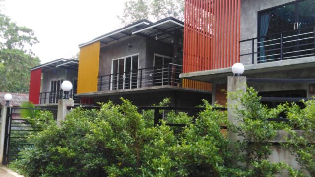 Modern gray two-story house with red and yellow vertical panels, balcony railings, and green bushes in front.