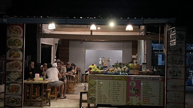 Small group of people dine at a brightly lit open-air street food stall at night, with menus and fresh fruits visible.