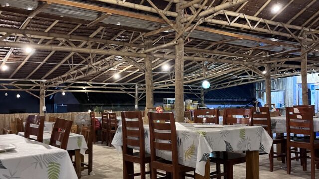 An open-air restaurant with wooden tables and chairs, white leaf-patterned tablecloths, and a bamboo roof at night.